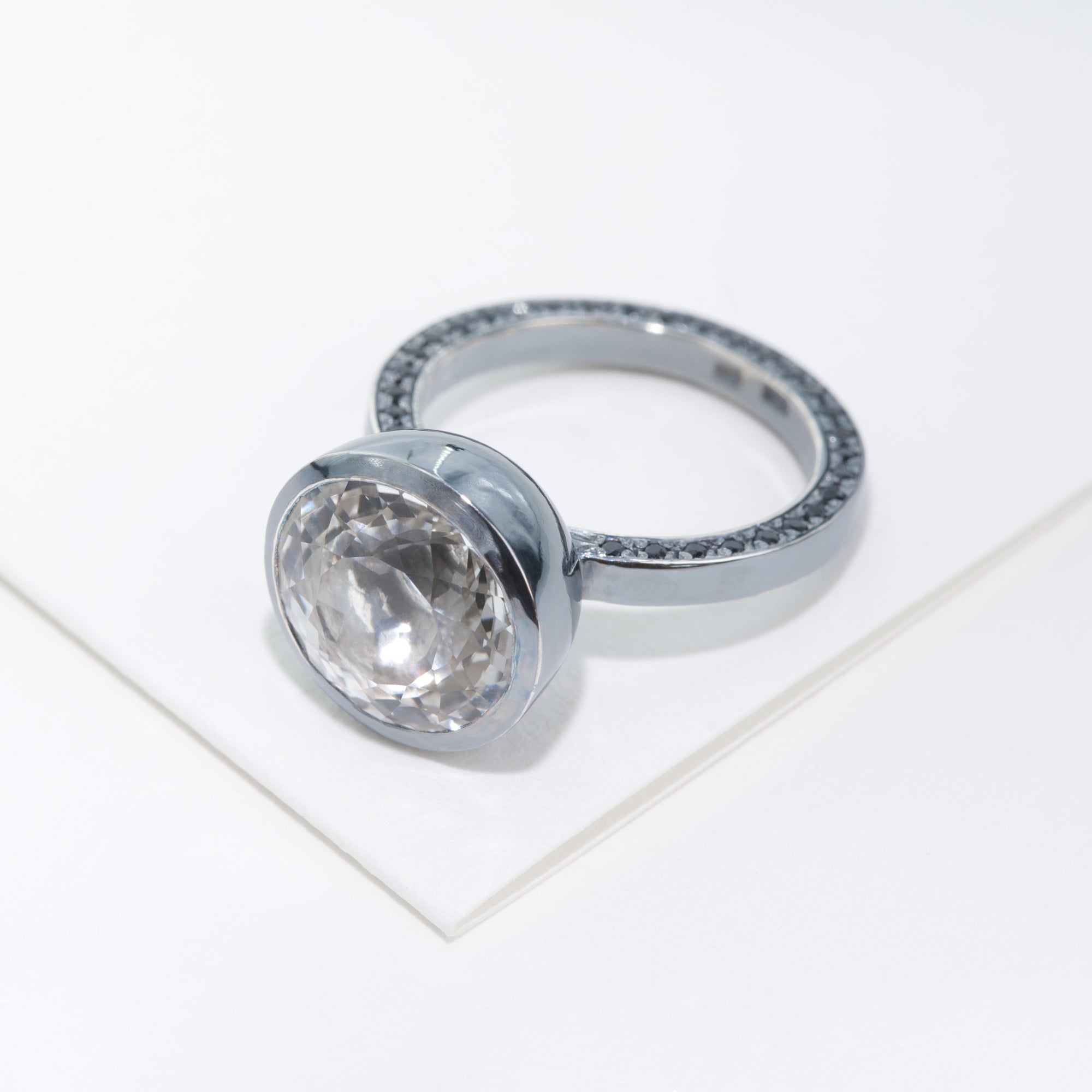 The ring with huge white stone as a focal and shank set with black stones is placed on white cardboard.