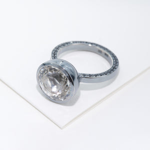 The ring with huge white stone as a focal and shank set with black stones is placed on white cardboard.