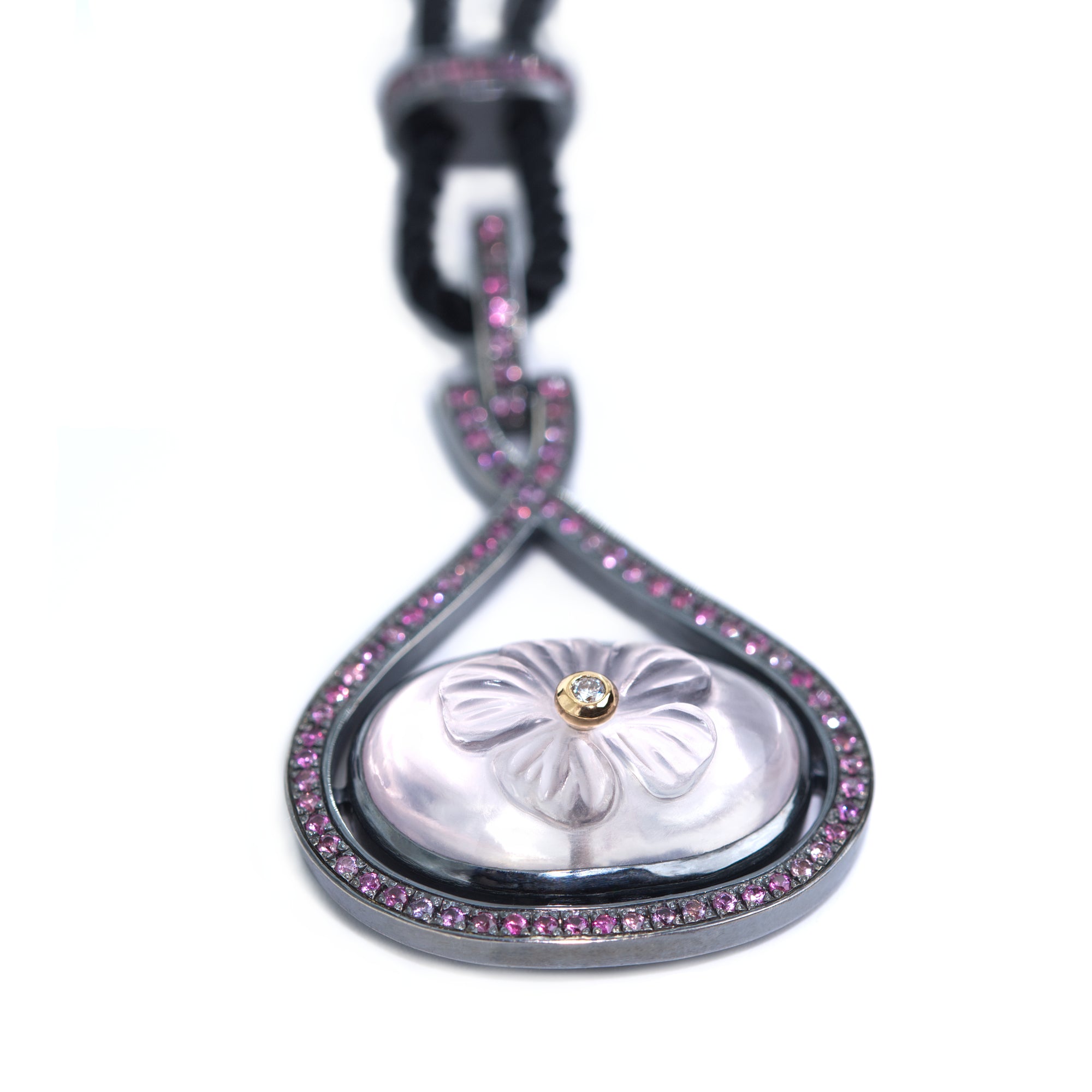 A necklace featuring carved rose quartz adorned with white diamond hangs from black rope. The necklace is presented on white background.