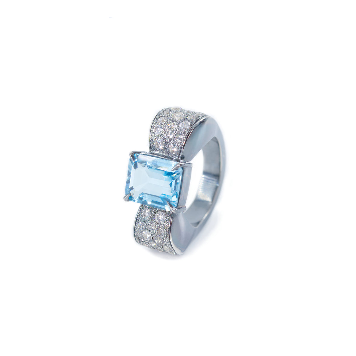 The photo shows the front view of the bow-shaped ring with the side of the ring slightly visible. The ring is placed vertically. The background of the photo is a white sheet of paper. In the center of the bow there is an octagonal blue topaz, the side of the bow is decorated with round white zircons. The ring is in a classic art deco style.