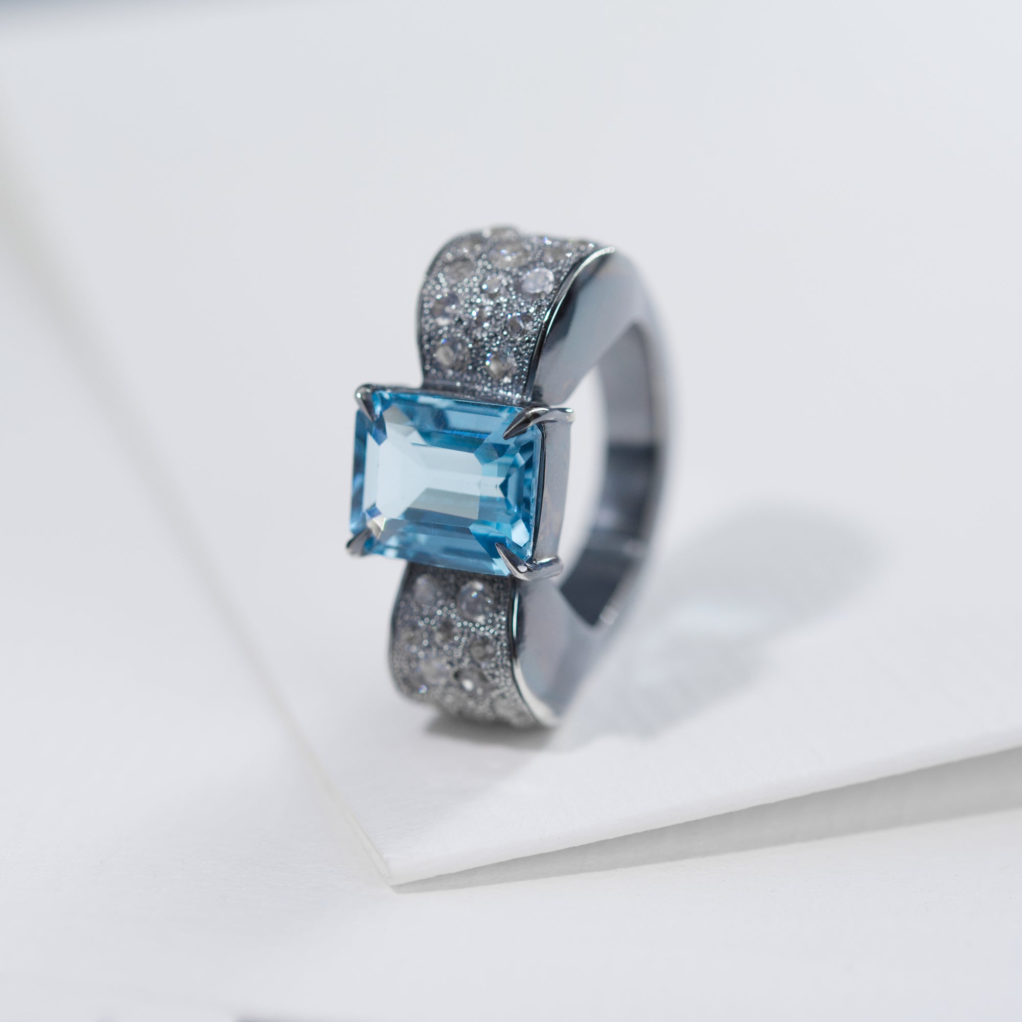 The photo shows the front view of the bow-shaped ring. The ring is placed vertically. The background of the photo is a white sheet of paper. In the center of the bow there is an octagonal blue topaz, the side of the bow is decorated with round white zircons. The ring is in a classic art deco style.