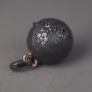 An orb locket placed on side is pictured on grey background. The medallion is adorned with hand carved cherry blossoms and pink sapphires. Piece by Ewa Z. Sleziona Jewellery