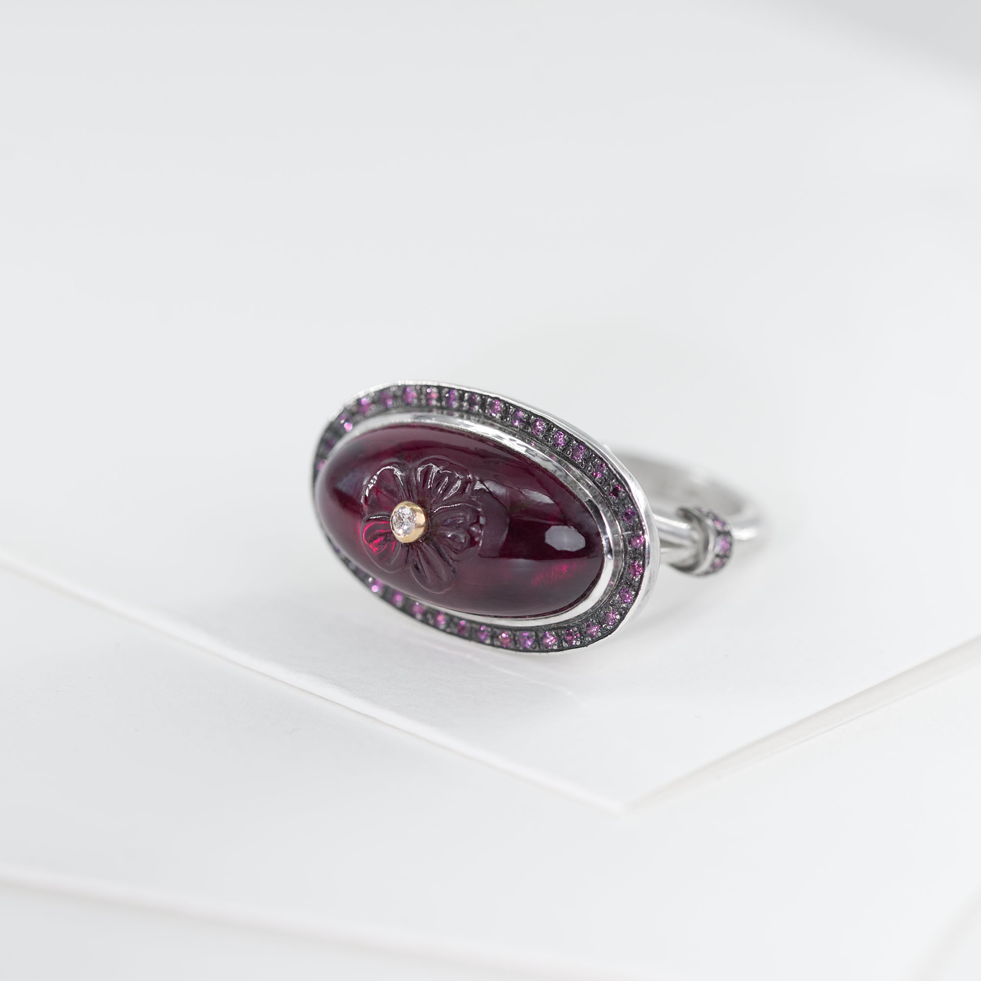Sterling silver ring with oval rhodolite cabochon. The gemstone is hand carved with flower and adorned with white diamond set in 18K gold. The ring's crown is finished with halo of rhodolites.