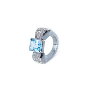 The photo shows the front view of the bow-shaped ring with the side of the ring slightly visible. The ring is placed vertically. The background of the photo is a white sheet of paper. In the center of the bow there is an octagonal blue topaz, the side of the bow is decorated with round white zircons. The ring is in a classic art deco style.