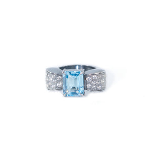 The photo shows the front view of the bow-shaped ring. The ring is placed horizontally. In the center of the bow there is an octagonal blue topaz, the side of the bow is decorated with round white zircons. The ring is in a classic art deco style.