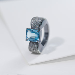 The photo shows the front view of the bow-shaped ring. The ring is placed vertically. The background of the photo is a white sheet of paper. In the center of the bow there is an octagonal blue topaz, the side of the bow is decorated with round white zircons. The ring is in a classic art deco style.