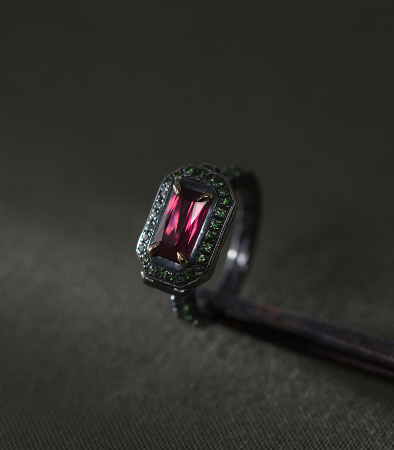 A blackened silver ring set with a rectangular rhodolite garnet held in four gold prongs, photographed close against a dark grey-green background. The stone is deep raspberry-red and lit from the front so the step cuts show as vertical bands of light. An octagonal frame of small green tsavorites surrounds it, and more tsavorites continue down both shoulders of the band. The metal is dark enough that the green stones read as the outline of the piece.