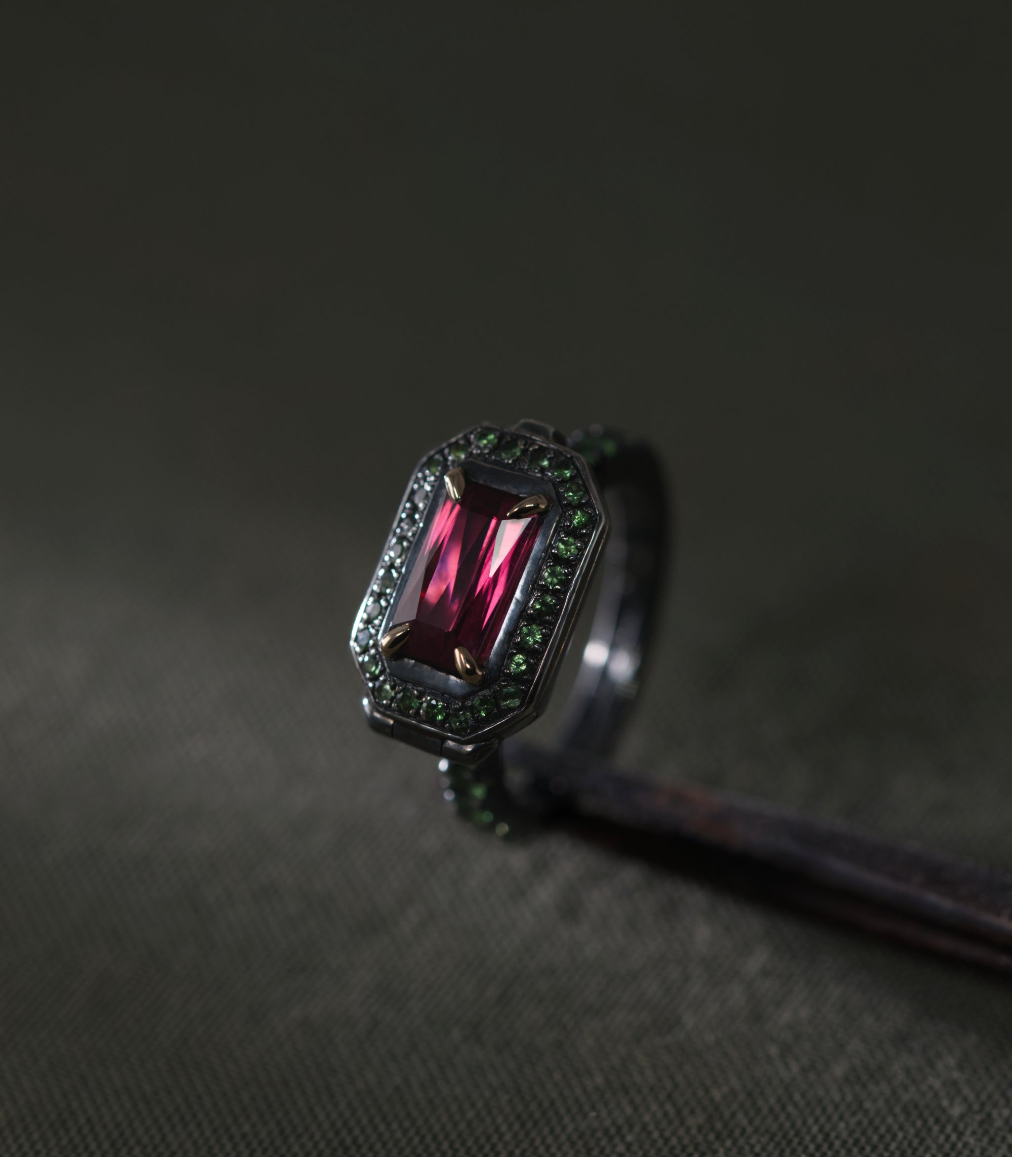 A blackened silver ring set with a rectangular rhodolite garnet held in four gold prongs, photographed close against a dark grey-green background. The stone is deep raspberry-red and lit from the front so the step cuts show as vertical bands of light. An octagonal frame of small green tsavorites surrounds it, and more tsavorites continue down both shoulders of the band. The metal is dark enough that the green stones read as the outline of the piece.