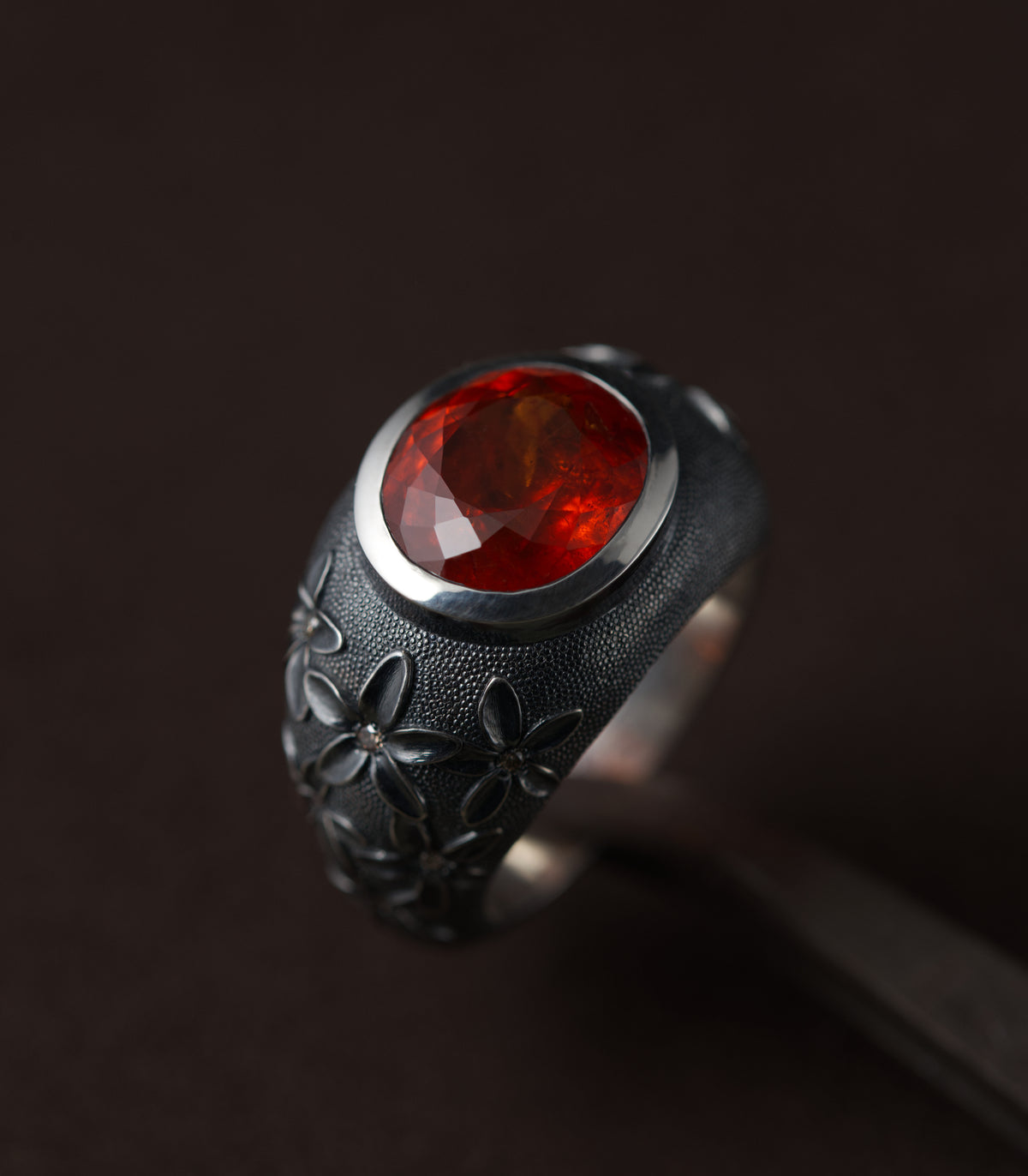 A silver ring with a domed, blackened band, set with a large oval spessartite garnet in a bright polished bezel. The stone is deep orange-red and lit from the front so the facets glow against the dark metal. Blossoms in relief climb the shoulders of the ring, each with a small brown diamond at its centre, and the surface between them is covered in fine hand-punched dots. The background is dark brown.