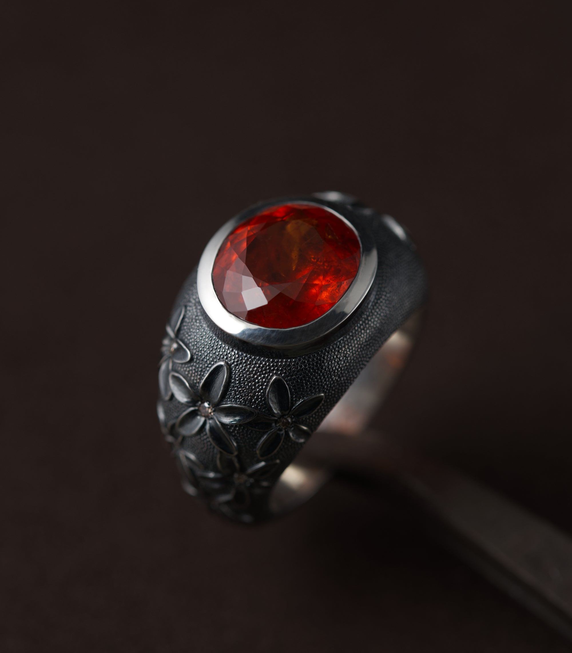 A silver ring with a domed, blackened band, set with a large oval spessartite garnet in a bright polished bezel. The stone is deep orange-red and lit from the front so the facets glow against the dark metal. Blossoms in relief climb the shoulders of the ring, each with a small brown diamond at its centre, and the surface between them is covered in fine hand-punched dots. The background is dark brown.