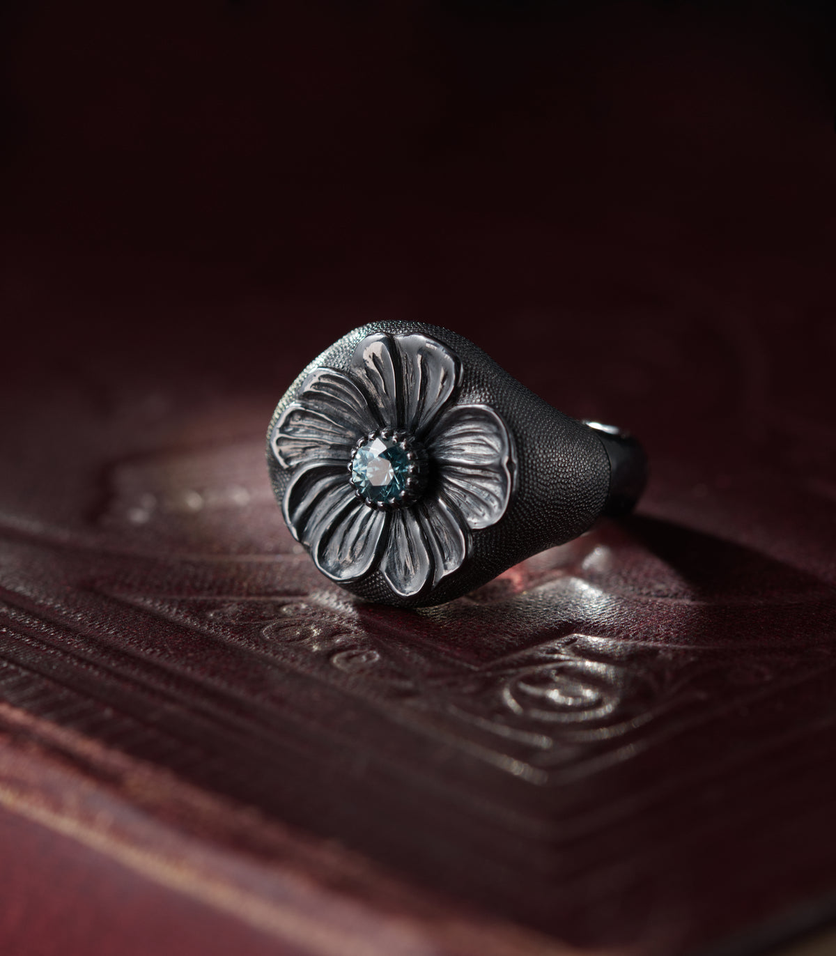 A heavy silver ring with a domed round head, photographed against dark red leather. A six-petalled flower is carved in relief across the face, its petals deeply grooved and polished bright against the blackened metal around them. At the centre sits a pale blue zircon in a claw setting. The ground surrounding the flower and the shoulders of the ring is covered in fine hand-punched dots.