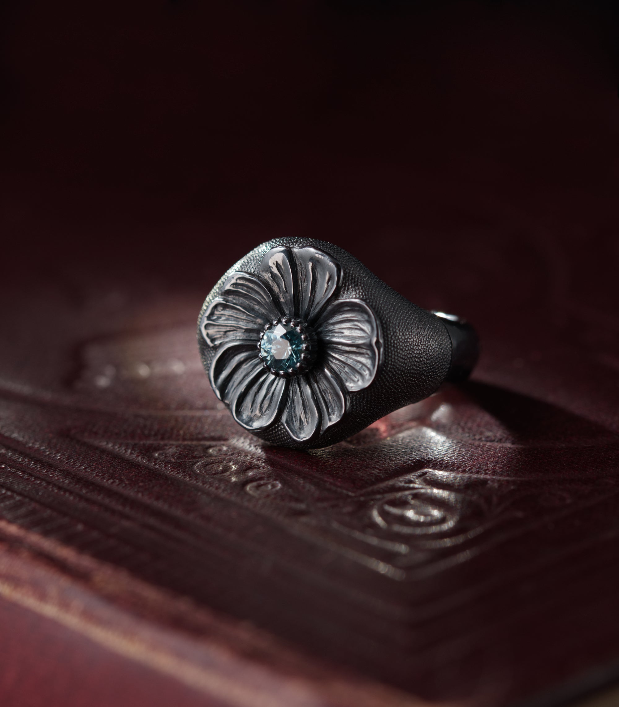 A heavy silver ring with a domed round head, photographed against dark red leather. A six-petalled flower is carved in relief across the face, its petals deeply grooved and polished bright against the blackened metal around them. At the centre sits a pale blue zircon in a claw setting. The ground surrounding the flower and the shoulders of the ring is covered in fine hand-punched dots.