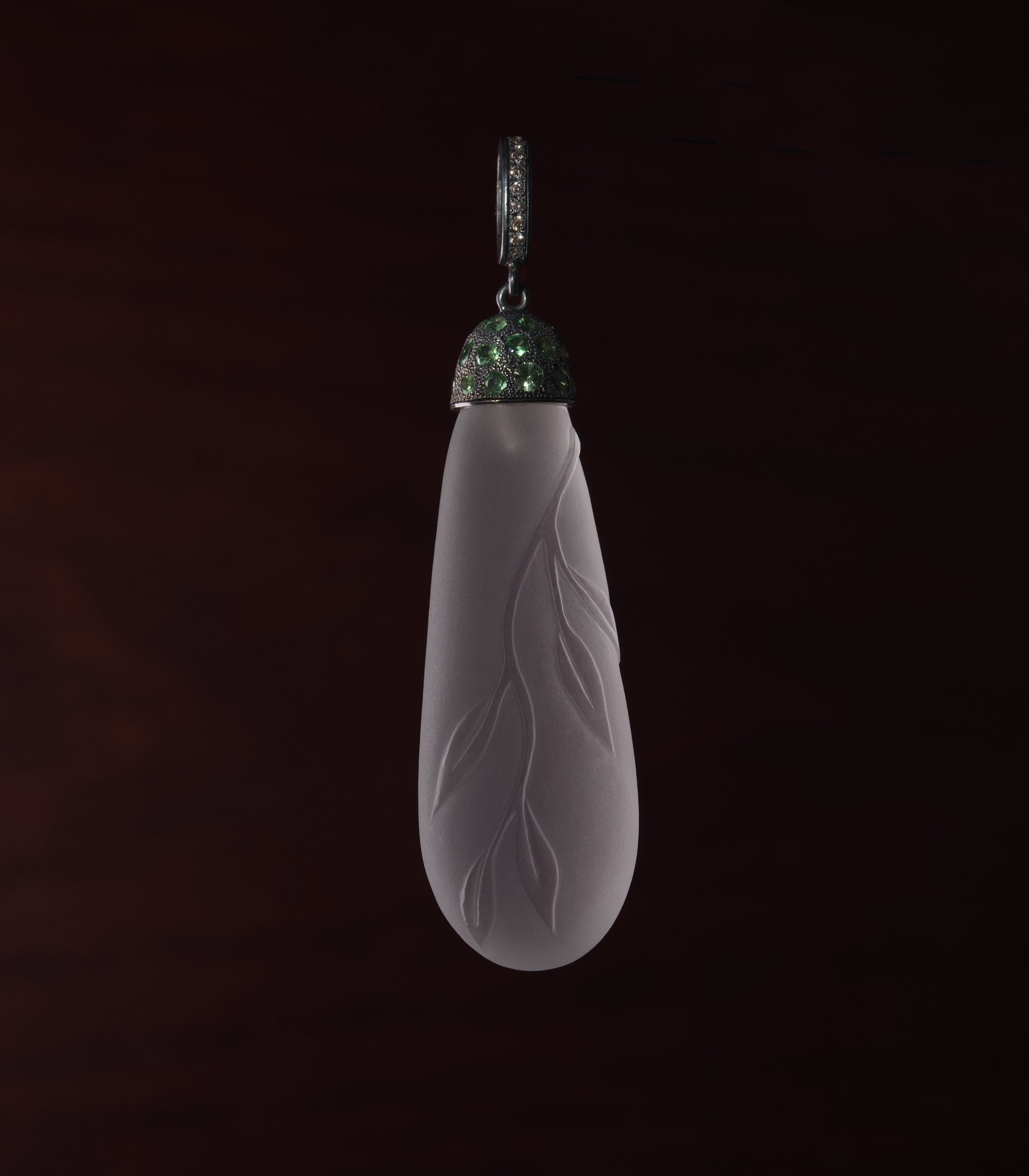 A long teardrop of frosted rock crystal hanging against a dark red-brown background. Leaves and a thin stem are carved in low relief across the front of the stone, rising from the matte surface. The cap at the top is blackened silver set closely with small green tsavorites, and the bail above it is set with a row of brown diamonds. The stone is lit from the front so the carving reads as shadow rather than shine.