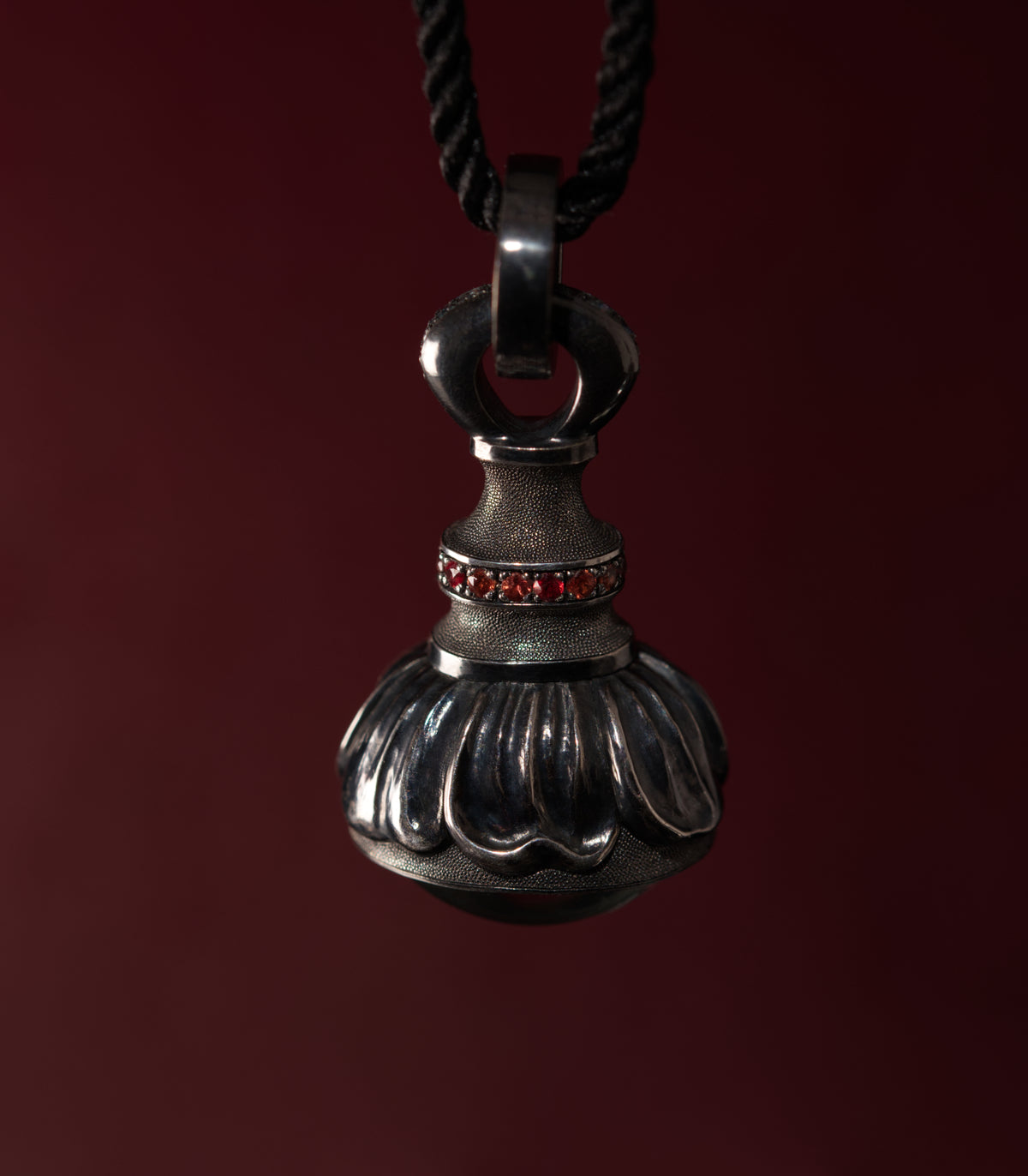 A silver pendant hanging from a black cord against a deep burgundy background, shaped like a small sealed vessel. The lower half is a heavy fluted dome with wide rounded lobes, and above it a narrow waist rises to a large hinged loop. A band of small red sapphires runs around the collar between the two. The narrow sections are covered in fine hand-punched dots, and the raised lobes are polished bright against the darkened metal.