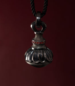 A silver pendant hanging from a black cord against a deep burgundy background, shaped like a small sealed vessel. The lower half is a heavy fluted dome with wide rounded lobes, and above it a narrow waist rises to a large hinged loop. A band of small red sapphires runs around the collar between the two. The narrow sections are covered in fine hand-punched dots, and the raised lobes are polished bright against the darkened metal.
