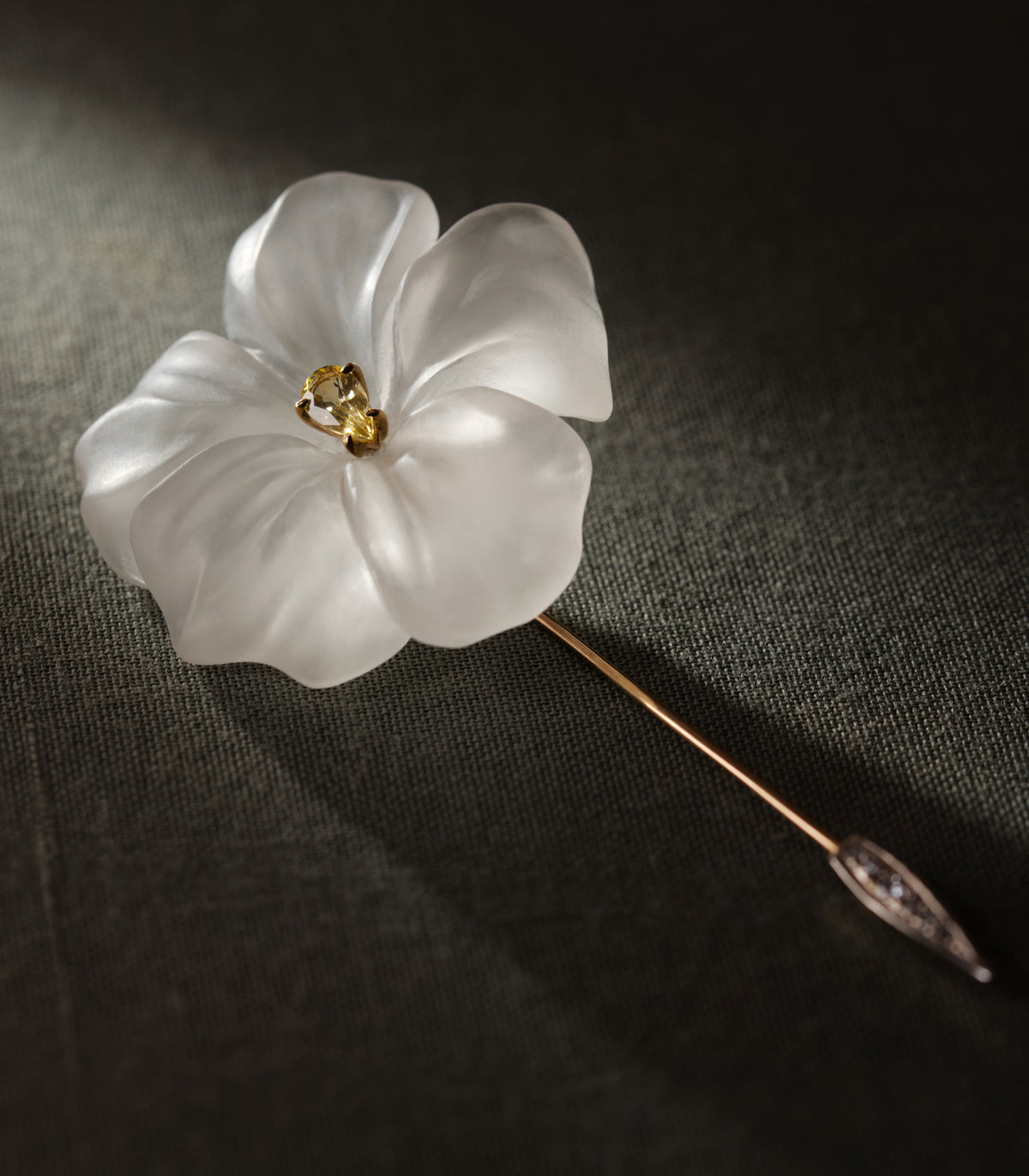 A flower carved from frosted rock crystal, lying at an angle on dark olive linen. Five petals curve outward from a centre set with a single yellow sapphire in gold. The long rose gold pin runs diagonally across the frame, widening near the point into a white gold spearhead set with small diamonds. The light falls from one side and passes through the stone rather than reflecting off it.