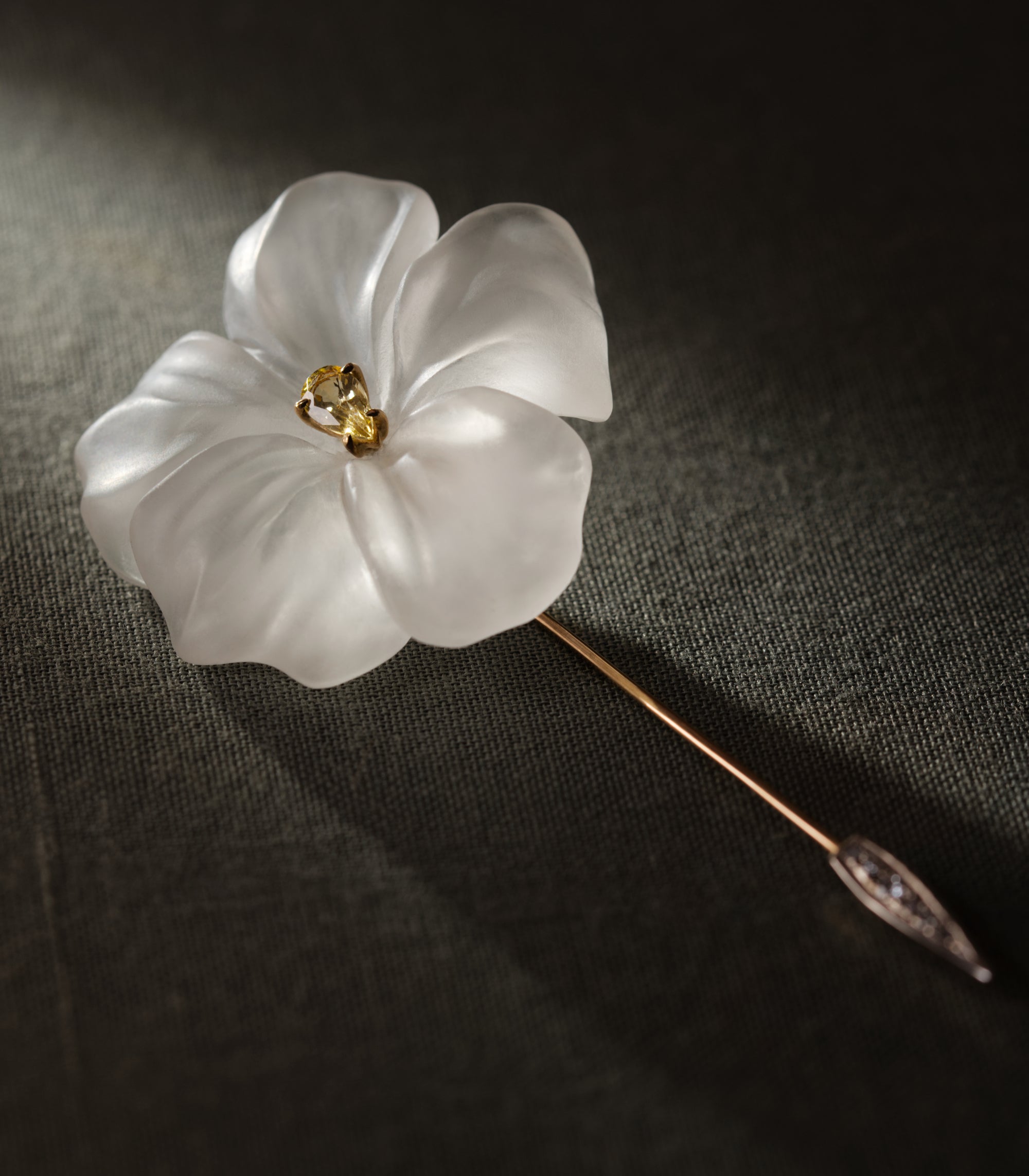 A flower carved from frosted rock crystal, lying at an angle on dark olive linen. Five petals curve outward from a centre set with a single yellow sapphire in gold. The long rose gold pin runs diagonally across the frame, widening near the point into a white gold spearhead set with small diamonds. The light falls from one side and passes through the stone rather than reflecting off it.