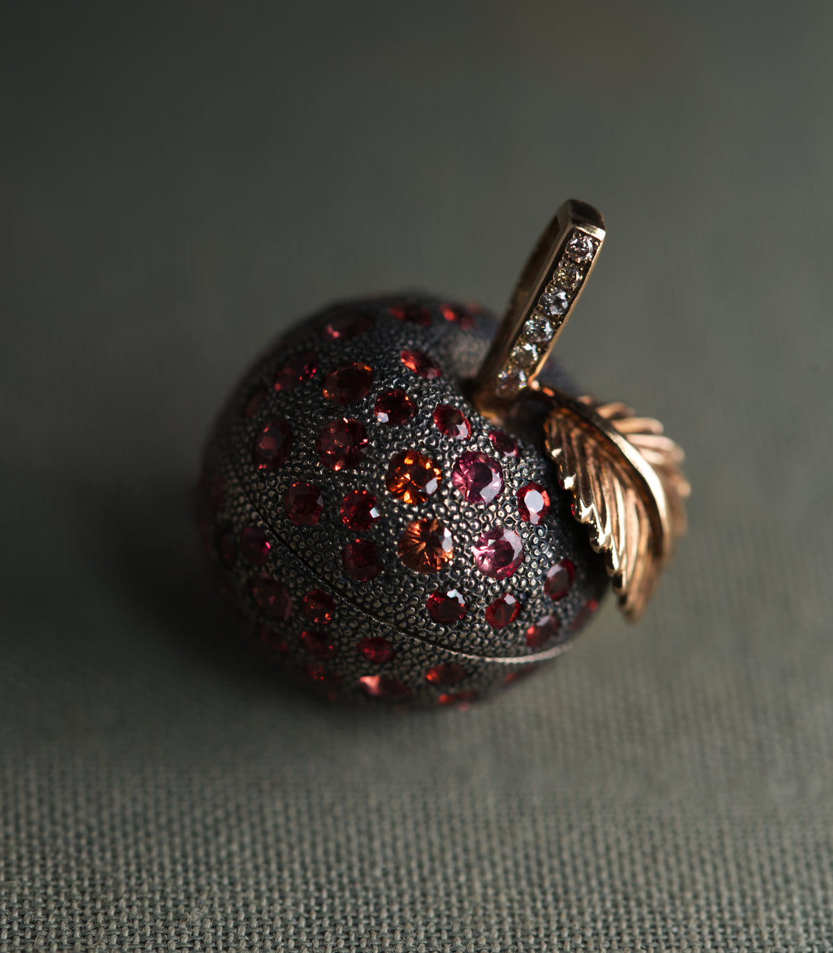 An apple-shaped locket in dark oxidised silver, photographed at close range against a grey-green background. The entire body is covered in fine hand-punched dots and set with round sapphires in red, raspberry and orange. A rose gold leaf curls away from the top, and the stem beside it is set with a row of brown diamonds.