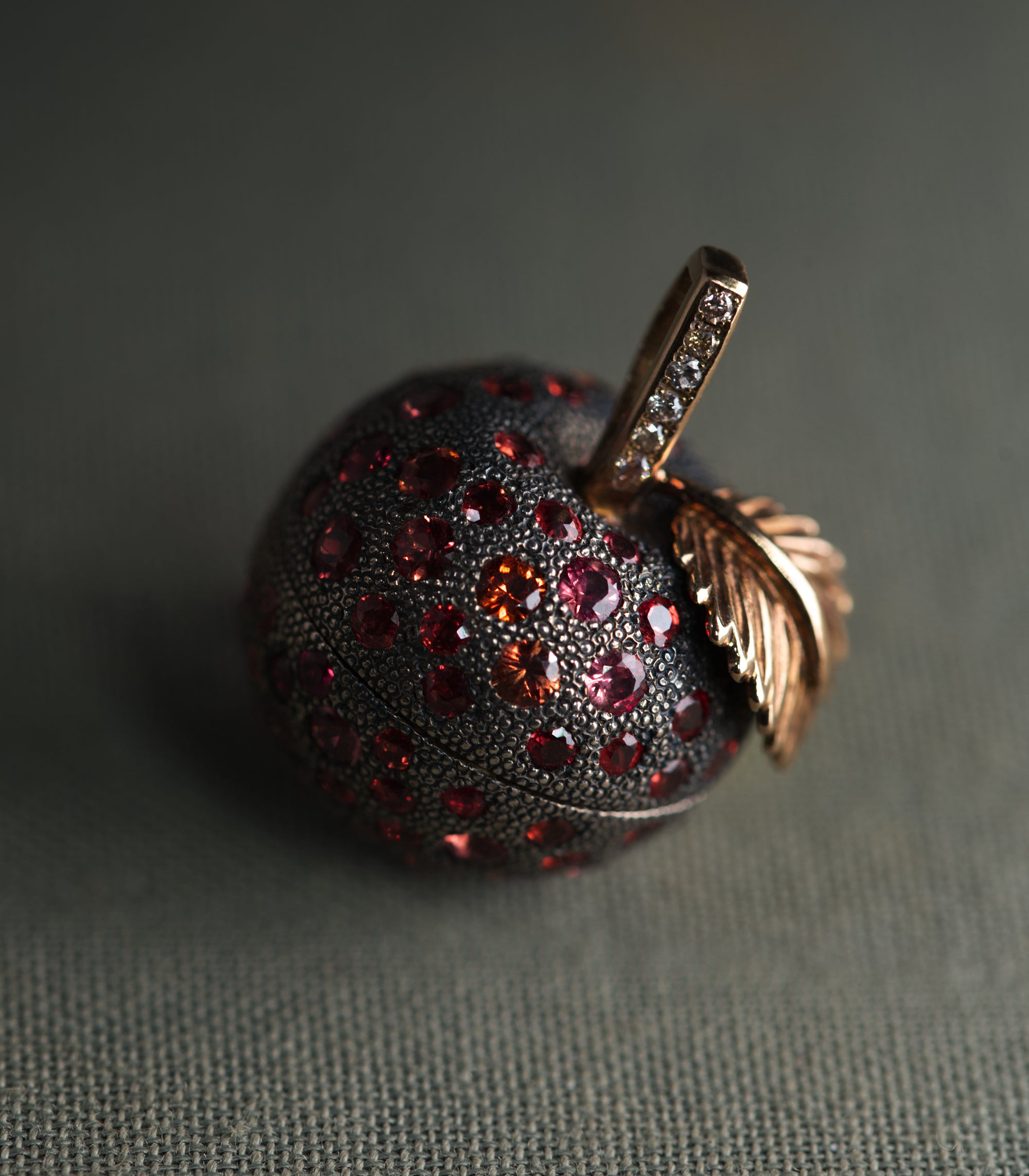 An apple-shaped locket in dark oxidised silver, photographed at close range against a grey-green background. The entire body is covered in fine hand-punched dots and set with round sapphires in red, raspberry and orange. A rose gold leaf curls away from the top, and the stem beside it is set with a row of brown diamonds.