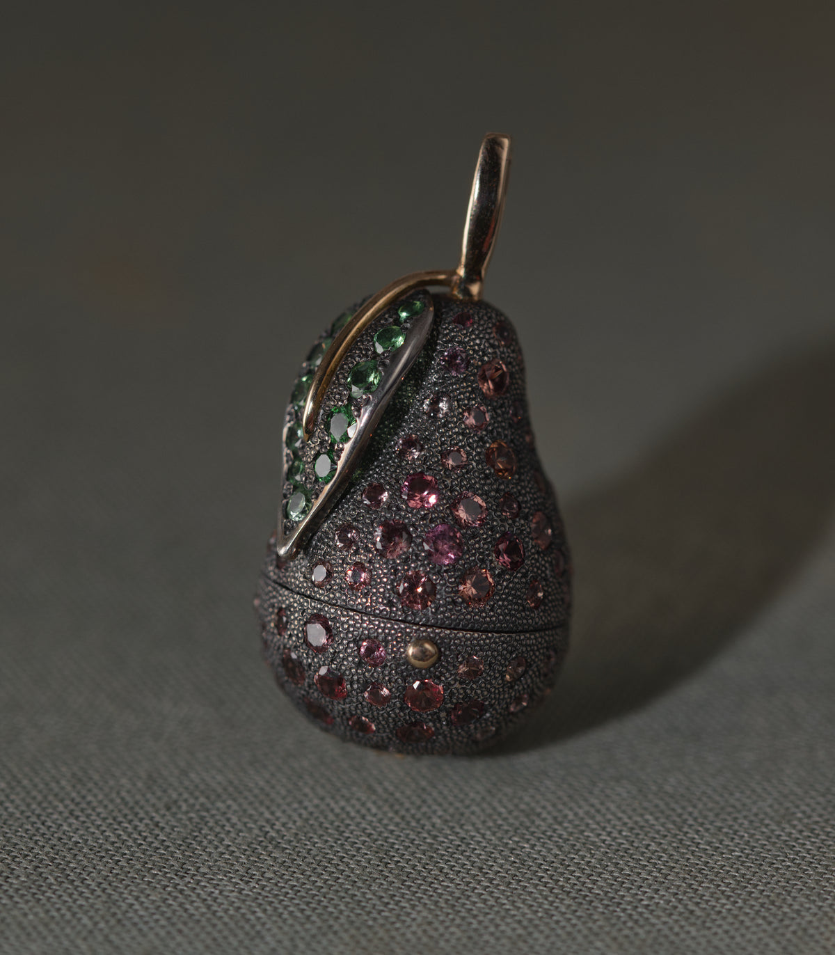 A pear-shaped locket in dark oxidised silver, photographed against a plain grey background. The whole body is covered in fine hand-punched dots and set with round sapphires running from rose to salmon. A silver leaf lies against the shoulder of the fruit, set with green tsavorites, its central vein picked out in gold. A gold stem rises from the top as the bail, and a fine seam runs around the lower third where the pear opens.