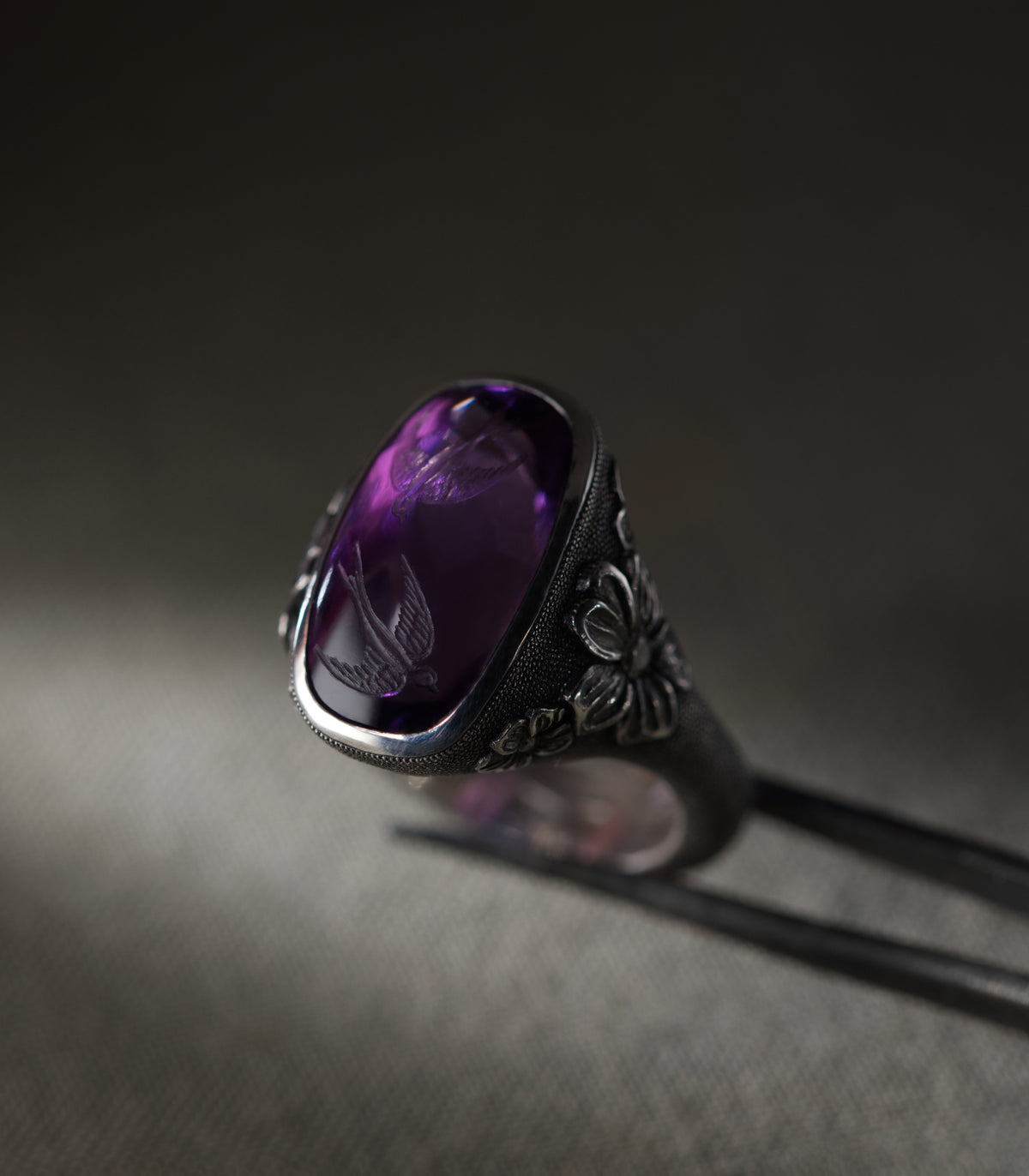 A silver ring set with a large cushion-shaped amethyst, photographed against a warm grey background and held in fine tweezers. Two swallows are carved into the face of the stone, and the curve of the cabochon carries the image down into the purple so that the birds appear to hang in the depth of it. Flowers in relief sit at the shoulders of the setting, each with a small diamond at its centre, and the surrounding metal is covered in fine hand-punched dots.