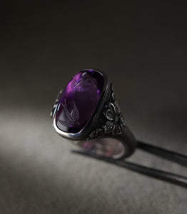 A silver ring set with a large cushion-shaped amethyst, photographed against a warm grey background and held in fine tweezers. Two swallows are carved into the face of the stone, and the curve of the cabochon carries the image down into the purple so that the birds appear to hang in the depth of it. Flowers in relief sit at the shoulders of the setting, each with a small diamond at its centre, and the surrounding metal is covered in fine hand-punched dots.