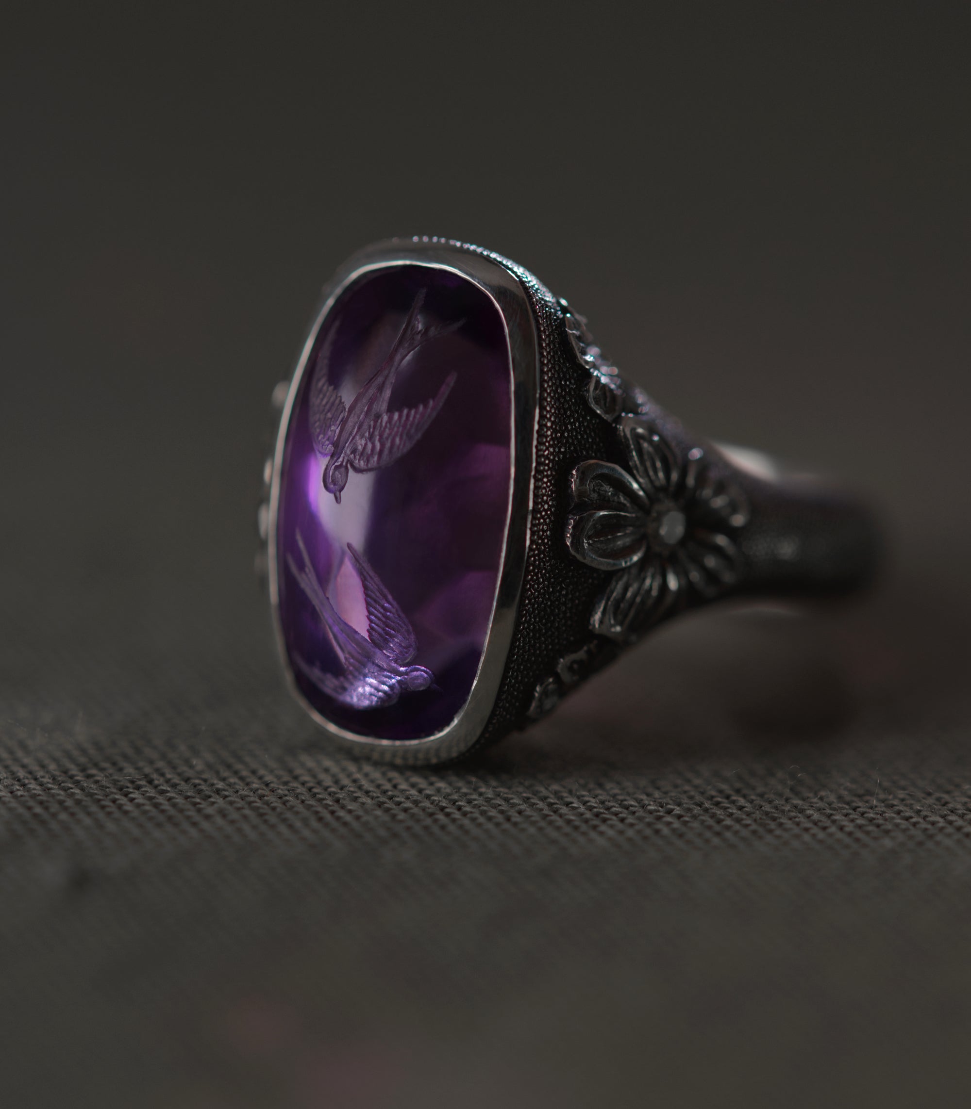 The ring seen from a low three-quarter angle against grey linen. The amethyst fills the upper left of the frame with both swallows visible in the depth of the stone, and the polished bezel throws a bright line along its edge. To the right, a flower carved in relief sits at the shoulder with a diamond at its centre, and the dotted ground runs down the shank and out of focus. The metal is dark throughout, the stone the only colour in the frame.