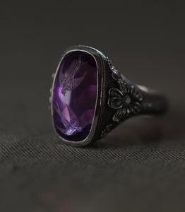 The ring seen from a low three-quarter angle against grey linen. The amethyst fills the upper left of the frame with both swallows visible in the depth of the stone, and the polished bezel throws a bright line along its edge. To the right, a flower carved in relief sits at the shoulder with a diamond at its centre, and the dotted ground runs down the shank and out of focus. The metal is dark throughout, the stone the only colour in the frame.