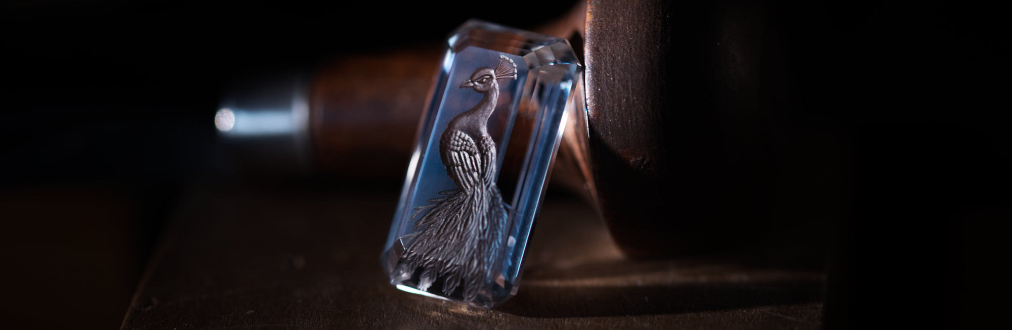 Rock crystal with carved intaglio presenting a peacock. The stone is faceted and bird's tail circles the facets. The graver is showed on the background.