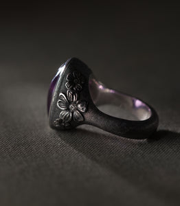 A silver ring seen from the side against grey linen, showing the depth of the setting and the shape of the shank. Three flowers of different sizes are carved in relief along the shoulder, each with a small diamond at its centre, standing out bright against metal darkened almost to black. The surface around them is covered in fine hand-punched dots. The inside of the band is left polished and catches a violet reflection from the stone above.