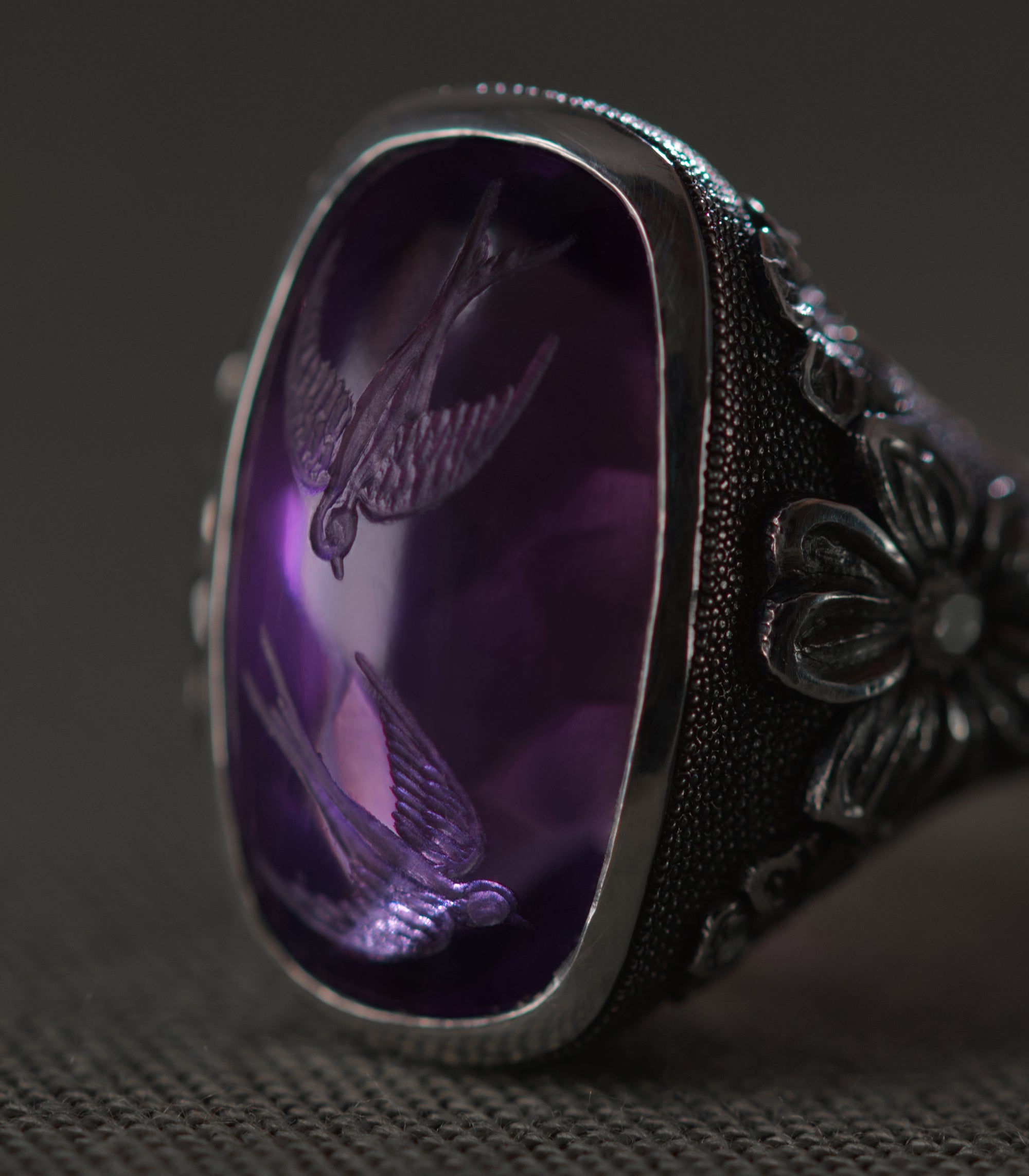 A close view of the amethyst from above, filling most of the frame. Two swallows are carved into the face of the stone, one above the other, wings open and tails spread as though in flight. The purple deepens toward the edges of the cabochon and the birds appear to hang inside it rather than on its surface. To the right, carved flowers and the dotted silver ground run out of focus toward the edge of the frame.