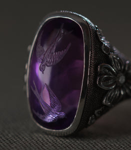 A close view of the amethyst from above, filling most of the frame. Two swallows are carved into the face of the stone, one above the other, wings open and tails spread as though in flight. The purple deepens toward the edges of the cabochon and the birds appear to hang inside it rather than on its surface. To the right, carved flowers and the dotted silver ground run out of focus toward the edge of the frame.